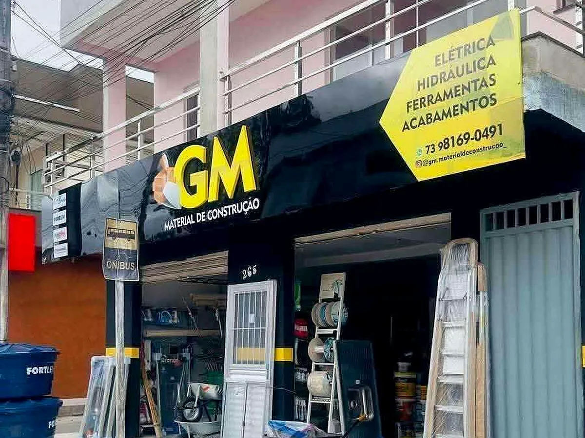 Fachada preta e amarela da GM Material de Construção, número 265 da Avenida Viana Gomes, com carrinhos de mão, escadas e caixas d'água na calçada